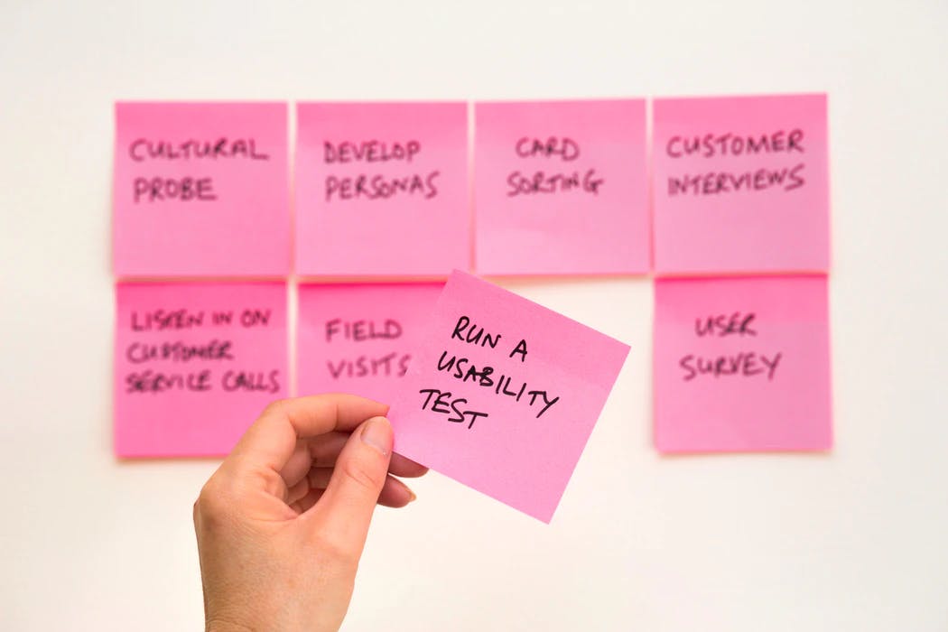 product development process steps written on pink post it notes with a hand pulling one off the wall that says run a usability test
